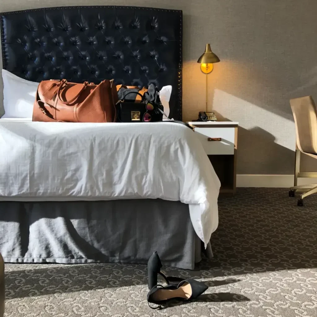 A sunlit hotel room featuring a made bed with a large, tufted navy headboard. A brown leather tote bag and a black purse sit on the white bedding.