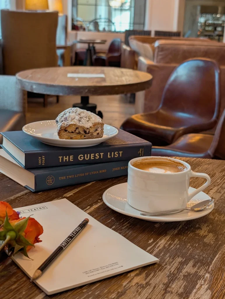 A cup of coffee, a scone on a stack of books, and a notepad sit on a wooden table in a cozy cafe or hotel lobby.