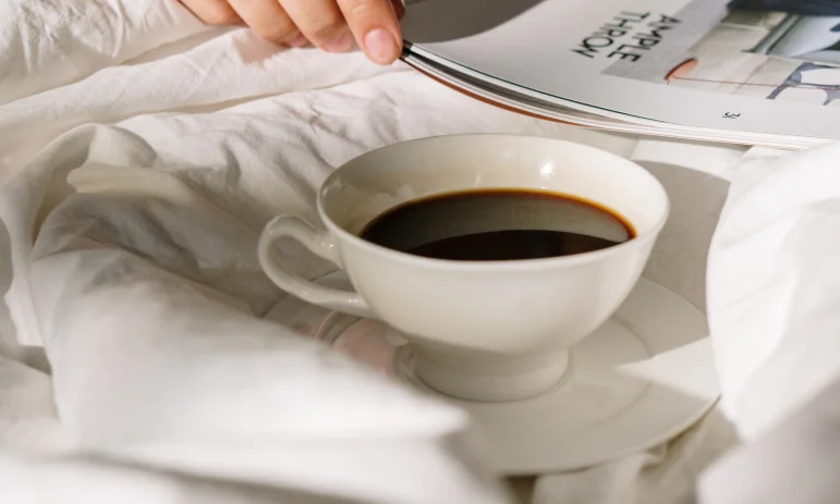 A close-up of a white cup of black coffee resting on a saucer amidst rumpled white bed sheets. In the background, a hand holds open a magazine, suggesting a relaxing morning.