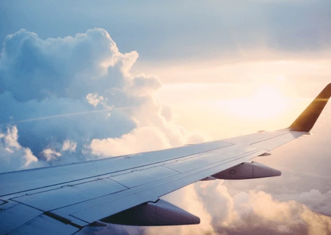 An airplane wing seen from a passenger window, soaring above a sea of fluffy clouds illuminated by the warm, golden light of a sunrise or sunset.