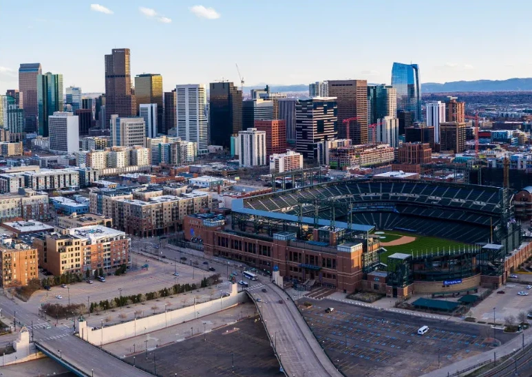 A daytime aerial photograph of downtown Denver, Colorado. In the foreground is the prominent brick structure of Coors Field, a large baseball stadium, surrounded by parking lots and city streets.