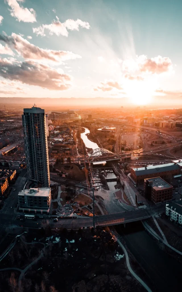 A dramatic aerial view of a city at sunset, with a tall skyscraper in the foreground as sunbeams radiate across the sky.