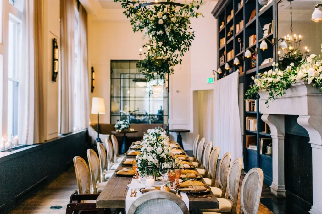 A long wooden table is set for an elegant dinner in a library-style room, with white floral centerpieces and a large floral arrangement hanging from the ceiling.