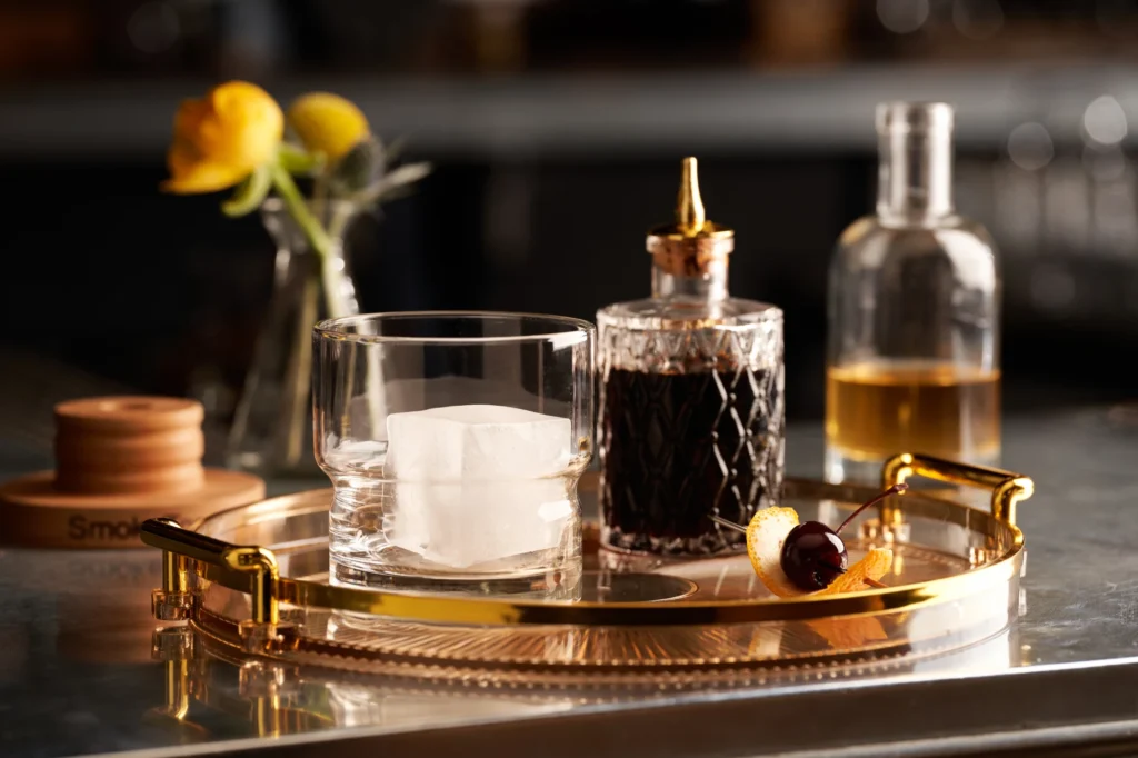 Ingredients for a classic cocktail are arranged on a gold tray, including a glass with a large ice cube, a bottle of bitters, and a garnish.