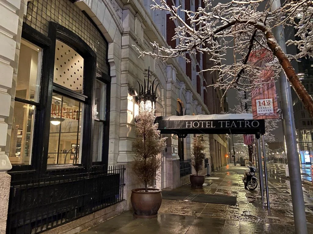 Snow-covered trees and the sleek entrance of Hotel Teatro illuminated by streetlights on a quiet, rainy night in Denver.
