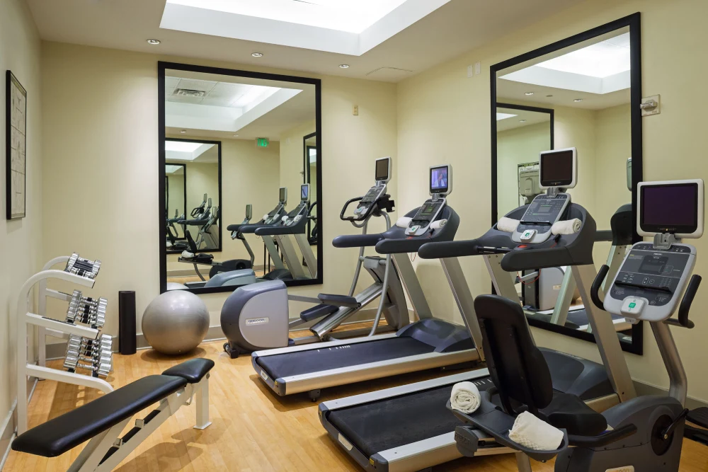 A bright, modern fitness center equipped with treadmills, an elliptical machine, a stationary bike, and a rack of dumbbells.