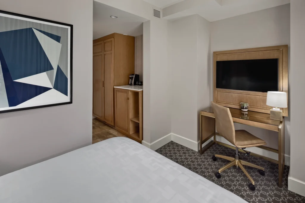 A hotel room features a bed in the foreground, a desk with a chair and lamp, a wall-mounted TV, a framed abstract artwork, and a small kitchenette area with a coffee maker.