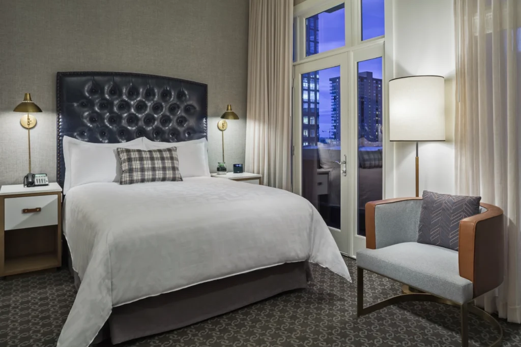 A well-appointed hotel room featuring a queen bed with a black tufted headboard, a modern armchair, and glass doors opening to a balcony with a city view at dusk.