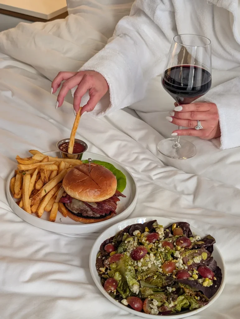 A person in a white bathrobe eats a meal of a burger, fries, and salad in bed while holding a glass of red wine.